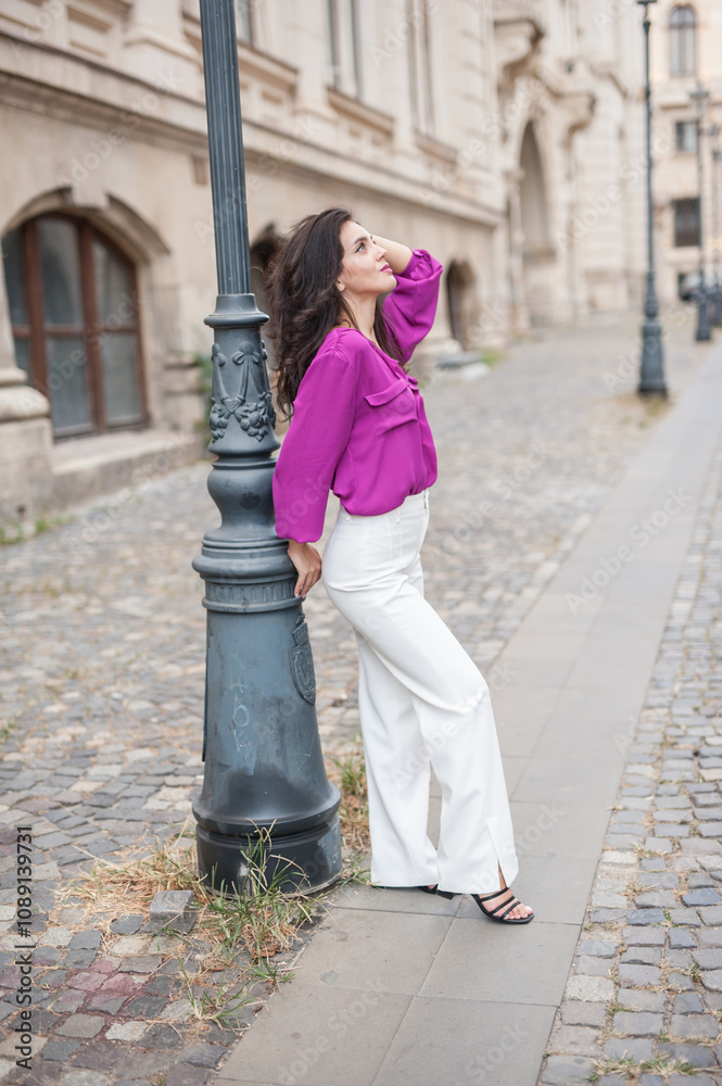 Beautiful young brunette woman in a purple blouse and white pants posing outdoor leans against a pillar .Portrait of a beautiful young brunette woman in a purple jacket posing