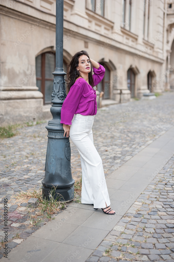 Beautiful young brunette woman in a purple blouse and white pants posing outdoor leans against a pillar .Portrait of a beautiful young brunette woman in a purple jacket posing