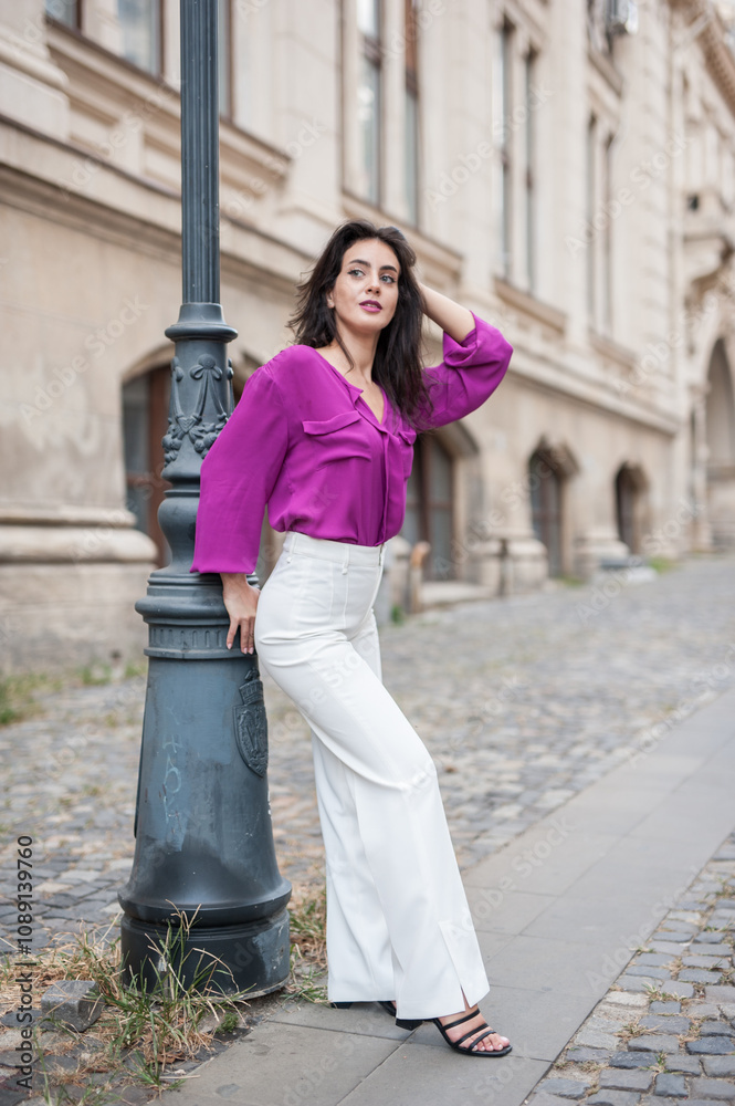 Beautiful young brunette woman in a purple blouse and white pants posing outdoor leans against a pillar .Portrait of a beautiful young brunette woman in a purple jacket posing