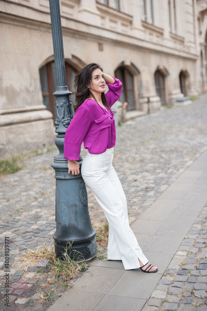 Beautiful young brunette woman in a purple blouse and white pants posing outdoor leans against a pillar .Portrait of a beautiful young brunette woman in a purple jacket posing
