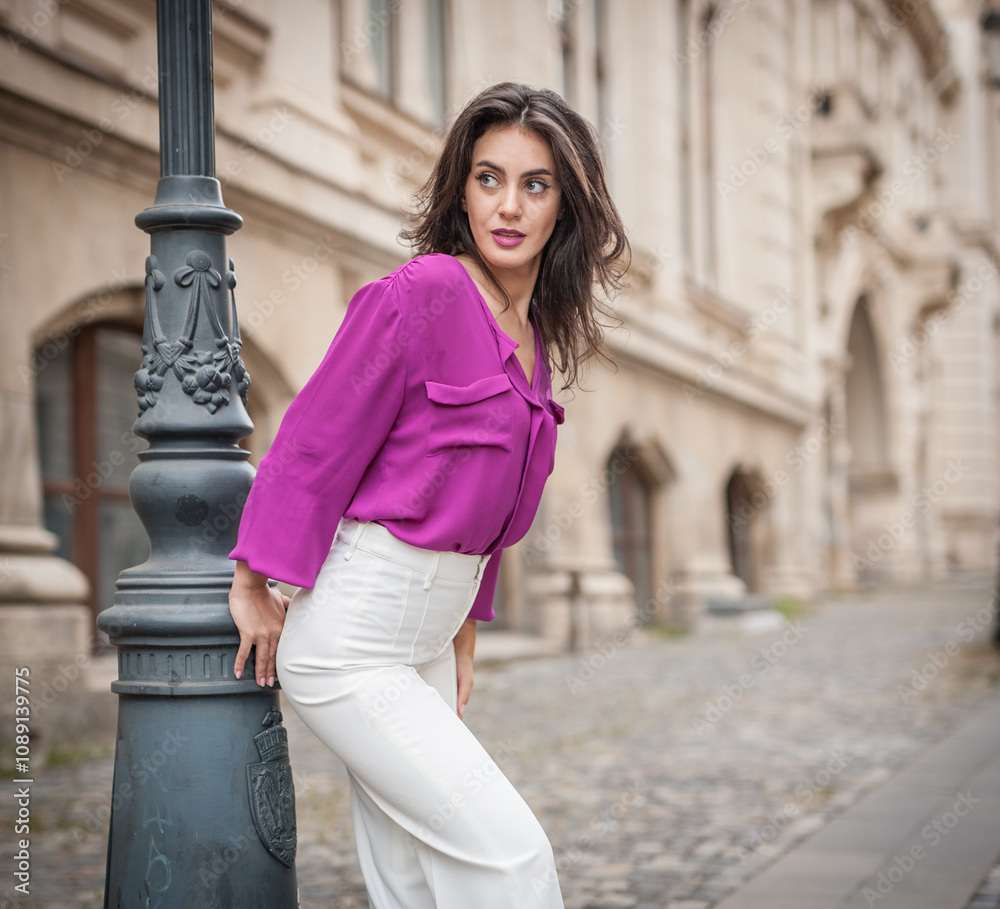 Beautiful young brunette woman in a purple blouse and white pants posing outdoor leans against a pillar .Portrait of a beautiful young brunette woman in a purple jacket posing