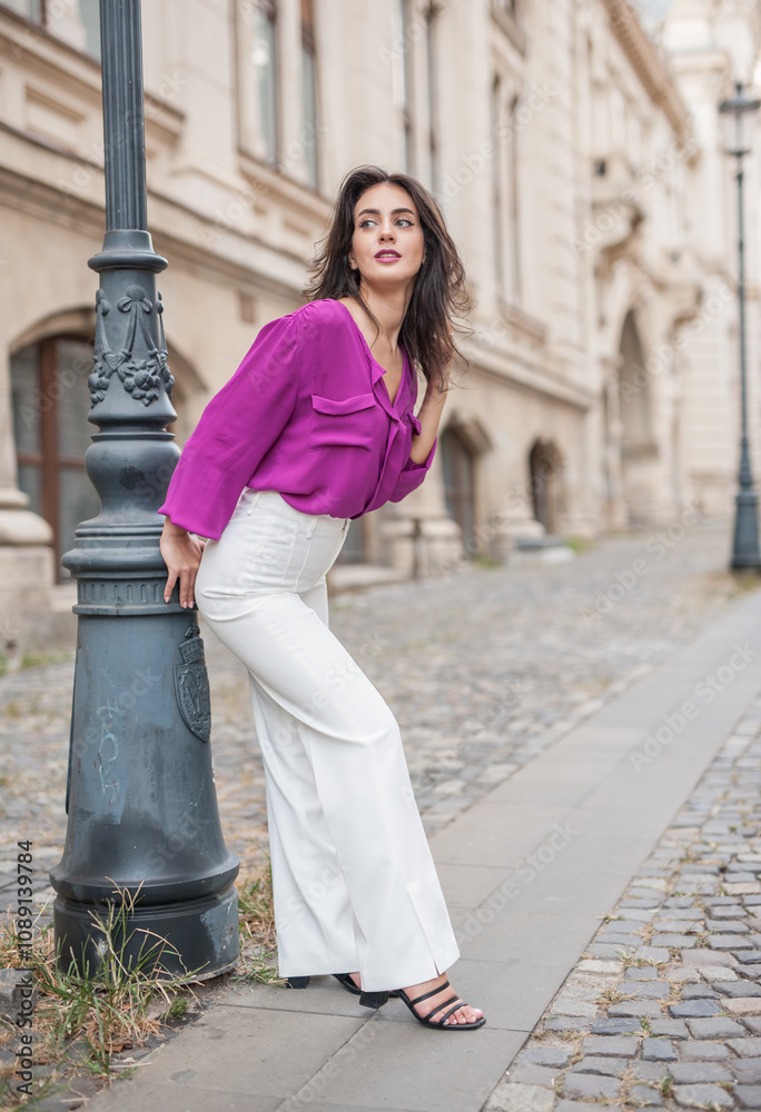 Beautiful young brunette woman in a purple blouse and white pants posing outdoor leans against a pillar .Portrait of a beautiful young brunette woman in a purple jacket posing