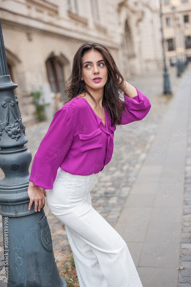 Beautiful young brunette woman in a purple blouse and white pants posing outdoor leans against a pillar .Portrait of a beautiful young brunette woman in a purple jacket posing
