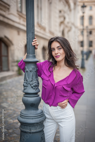 Obraz na płótnie Beautiful young brunette woman in a purple blouse and white pants posing outdoor leans against a pillar .Portrait of a beautiful young brunette woman in a purple jacket posing w salonie
