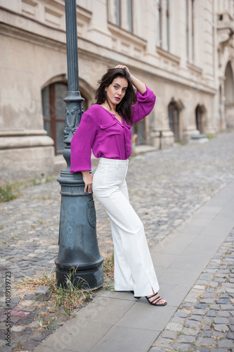 Obraz na płótnie Beautiful young brunette woman in a purple blouse and white pants posing outdoor leans against a pillar .Portrait of a beautiful young brunette woman in a purple jacket posing w salonie