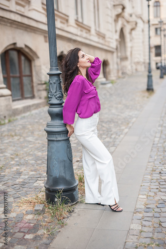 Obraz na płótnie Beautiful young brunette woman in a purple blouse and white pants posing outdoor leans against a pillar .Portrait of a beautiful young brunette woman in a purple jacket posing w salonie
