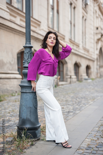 Obraz na płótnie Beautiful young brunette woman in a purple blouse and white pants posing outdoor leans against a pillar .Portrait of a beautiful young brunette woman in a purple jacket posing w salonie