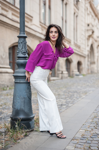 Obraz na płótnie Beautiful young brunette woman in a purple blouse and white pants posing outdoor leans against a pillar .Portrait of a beautiful young brunette woman in a purple jacket posing w salonie