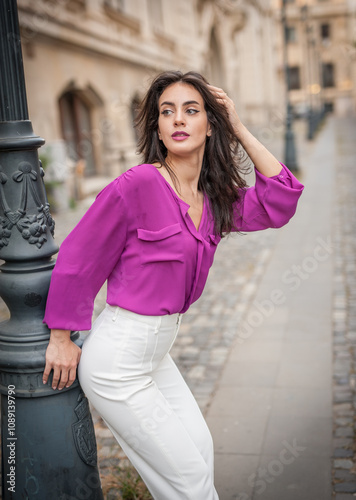 Obraz na płótnie Beautiful young brunette woman in a purple blouse and white pants posing outdoor leans against a pillar .Portrait of a beautiful young brunette woman in a purple jacket posing w salonie