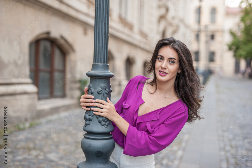 Beautiful young brunette woman in a purple blouse and white pants posing outdoor leans against a pillar .Portrait of a beautiful young brunette woman in a purple jacket posing