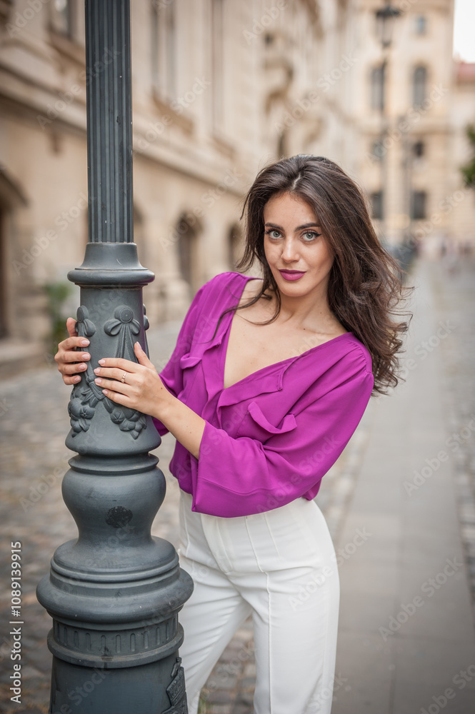 Beautiful young brunette woman in a purple blouse and white pants posing outdoor leans against a pillar .Portrait of a beautiful young brunette woman in a purple jacket posing