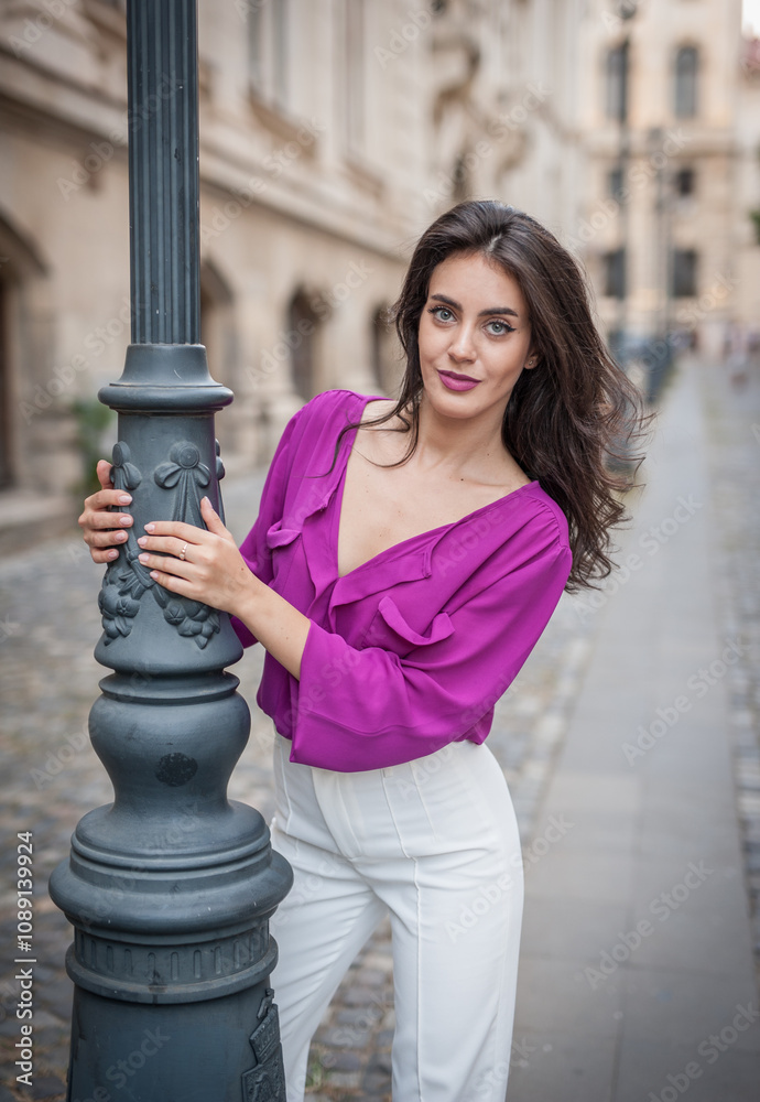 Beautiful young brunette woman in a purple blouse and white pants posing outdoor leans against a pillar .Portrait of a beautiful young brunette woman in a purple jacket posing