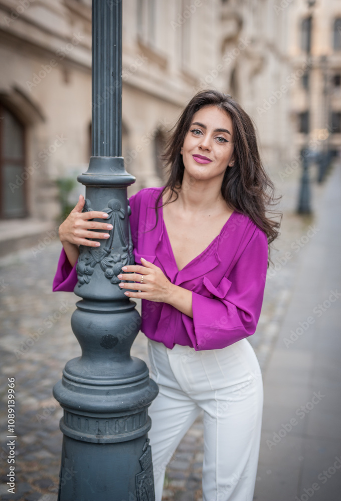 Beautiful young brunette woman in a purple blouse and white pants posing outdoor leans against a pillar .Portrait of a beautiful young brunette woman in a purple jacket posing