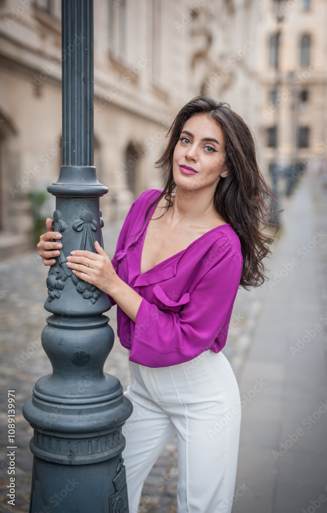 Beautiful young brunette woman in a purple blouse and white pants posing outdoor leans against a pillar .Portrait of a beautiful young brunette woman in a purple jacket posing