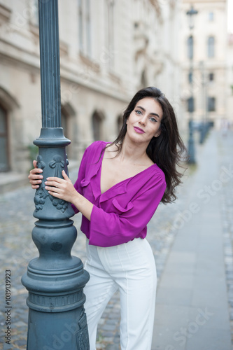 Beautiful young brunette woman in a purple blouse and white pants posing outdoor leans against a pillar .Portrait of a beautiful young brunette woman in a purple jacket posing 