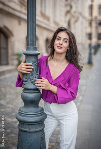 Obraz na płótnie Beautiful young brunette woman in a purple blouse and white pants posing outdoor leans against a pillar .Portrait of a beautiful young brunette woman in a purple jacket posing w salonie