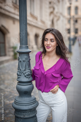 Obraz na płótnie Beautiful young brunette woman in a purple blouse and white pants posing outdoor leans against a pillar .Portrait of a beautiful young brunette woman in a purple jacket posing w salonie