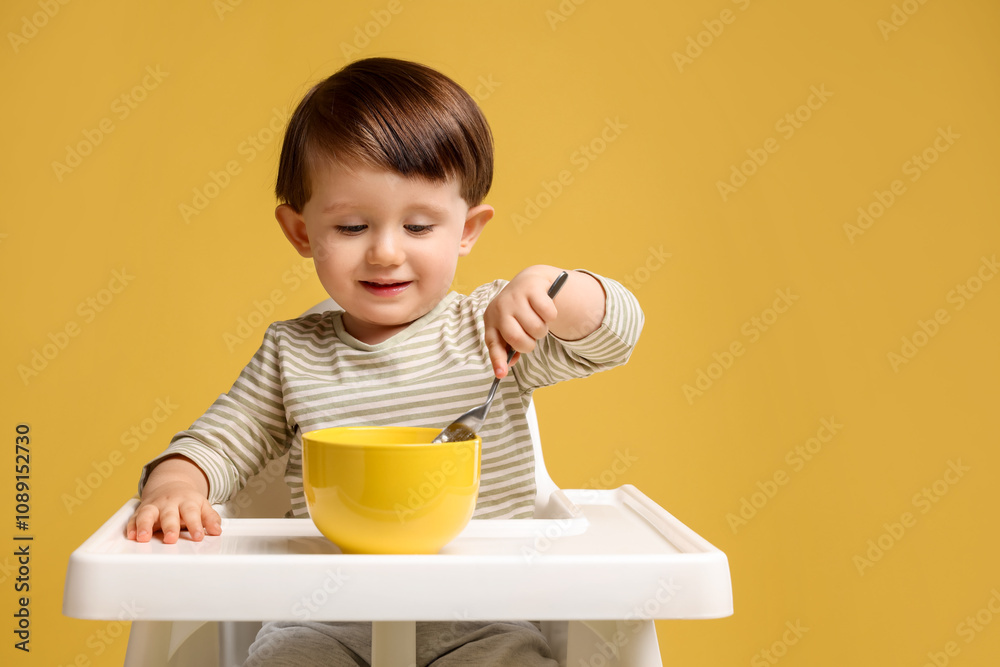 Cute little kid eating healthy baby food from bowl in high chair on yellow background Cute little kid eating healthy baby food from bowl in high chair on yellow background
