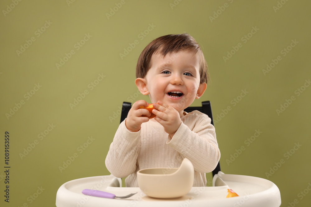 Cute little baby eating healthy food from bowl in high chair on olive background, space for text Cute little baby eating healthy food from bowl in high chair on olive background, space for text