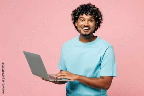 Young smiling happy IT Indian man he wear blue t-shirt casual clothes hold use work on laptop pc computer look camera isolated on plain pastel light pink background studio portrait. Lifestyle concept. photo