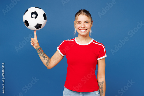 Young smiling happy cheerful fun woman fan wear basic red t-shirt cheer up support football sport team hold in hand spin soccer ball watch tv live stream look camera isolated on plain blue background.