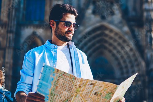 Bearded young man tourist in black sunglasses standing in front of old architectural building in downtown and holding map in hands to searching right route and destination of showplaces in new city photo