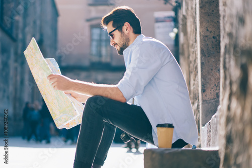 Pensive bearded tourist in sunglasses reading travel map with showplaces of architectural city sitting on stairs on street.Stylish male traveler searching right direction of destination during trip photo