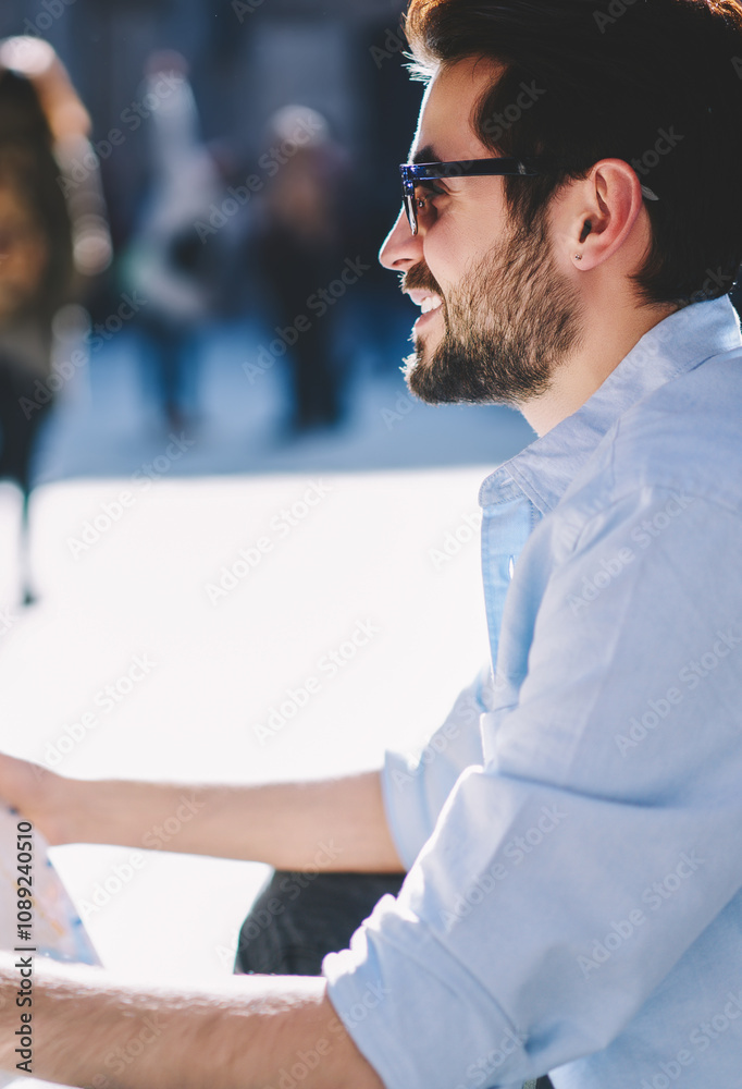 Fototapeta premium Cheerful bearded tourist in sunglasse resting outdoors on street of architectural downtown.Positive young man traveler in spectacles holding map in hands to find showplaces during trip