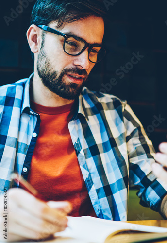 Pensive bearded student rewriting text information from smartphone device in notepad.Hipster guy in eyeglasses reading news on website on telephone and making notes in notebook sitting in coffee shop