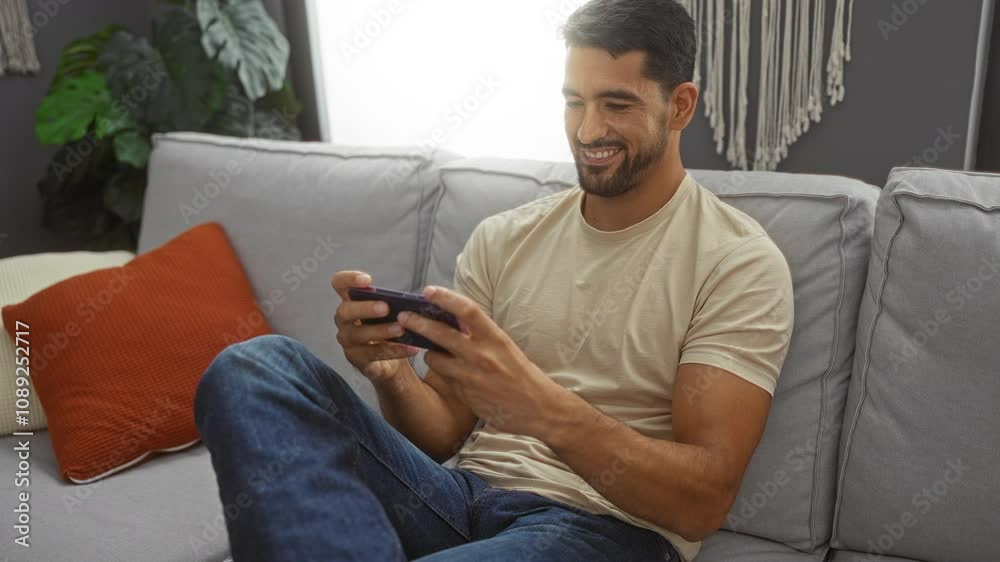 Young man gaming on smartphone in living room with gray couch and orange pillow, smiling and relaxed. Young man gaming on smartphone in living room with gray couch and orange pillow, smiling and relaxed.