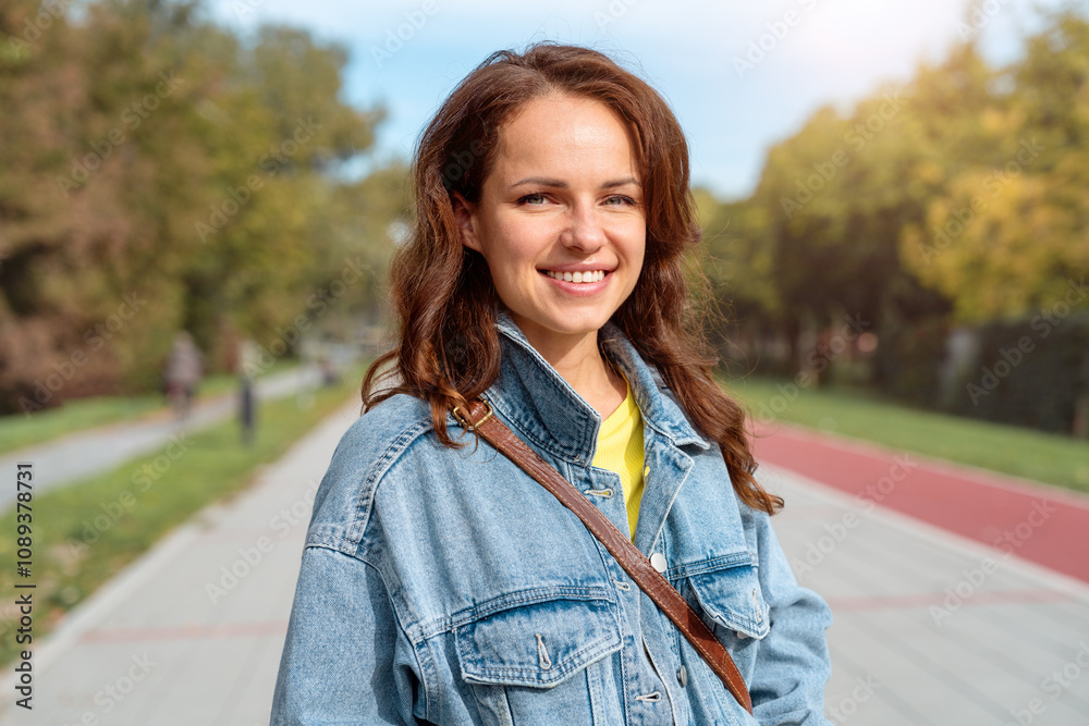Portrait of young brunette Caucasian woman in denim jacket walking outdoors, looking at camera and smiling. Portrait of young brunette Caucasian woman in denim jacket walking outdoors, looking at camera and smiling.