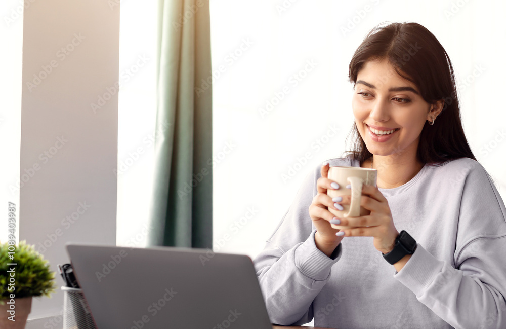People And Technology Concept. Portrait of smiling young woman using laptop and holding cup and drinking hot coffee. Female sitting at desk, watching video, movie or webinar, taking break People And Technology Concept. Portrait of smiling young woman using laptop and holding cup and drinking hot coffee. Female sitting at desk, watching video, movie or webinar, taking break
