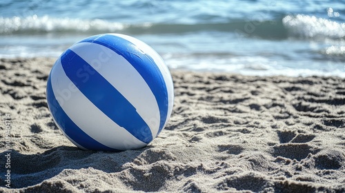 Colorful beach ball with blue and white stripes resting on sandy shore, gently lapped by tranquil ocean waves in soft natural light.