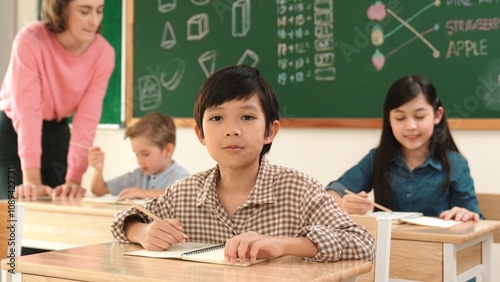 Asian smart boy smiling to camera while student writing answer in answer sheet. Multicultural student doing classwork or test while caucasian teacher checking student homework at classroom. Pedagogy.