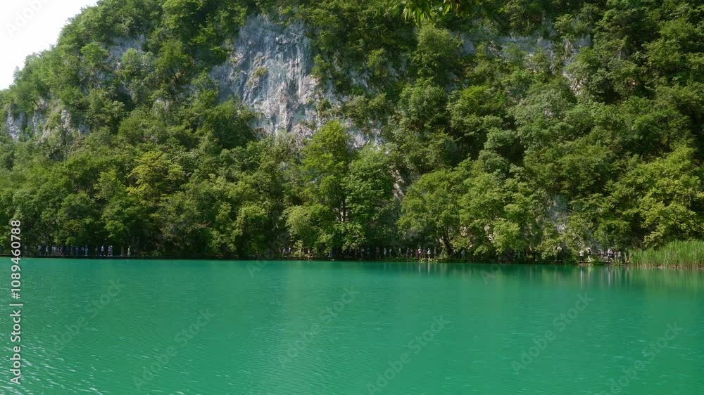 A crowd of tourists walking on the popular hiking trail near the cliffs by the shores of the Plitvice Lakes with clear azure water. The landmark of Croatia, included in the UNESCO World Heritage List. A crowd of tourists walking on the popular hiking trail near the cliffs by the shores of the Plitvice Lakes with clear azure water. The landmark of Croatia, included in the UNESCO World Heritage List.