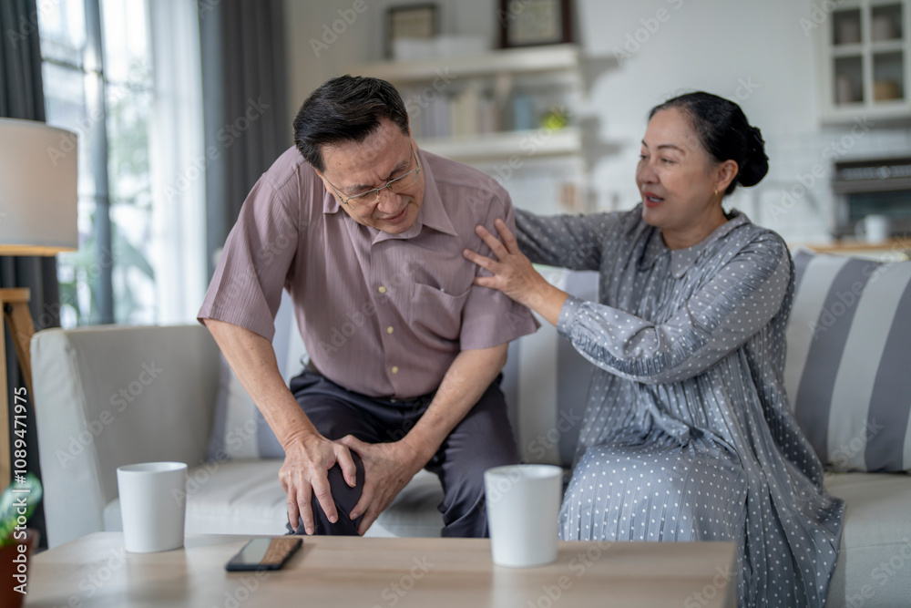 An elderly couple sits in their living room. The man holds his knee, appearing to be in discomfort, while his partner offers support. The image conveys care, concern, and the bond between the couple. An elderly couple sits in their living room. The man holds his knee, appearing to be in discomfort, while his partner offers support. The image conveys care, concern, and the bond between the couple.