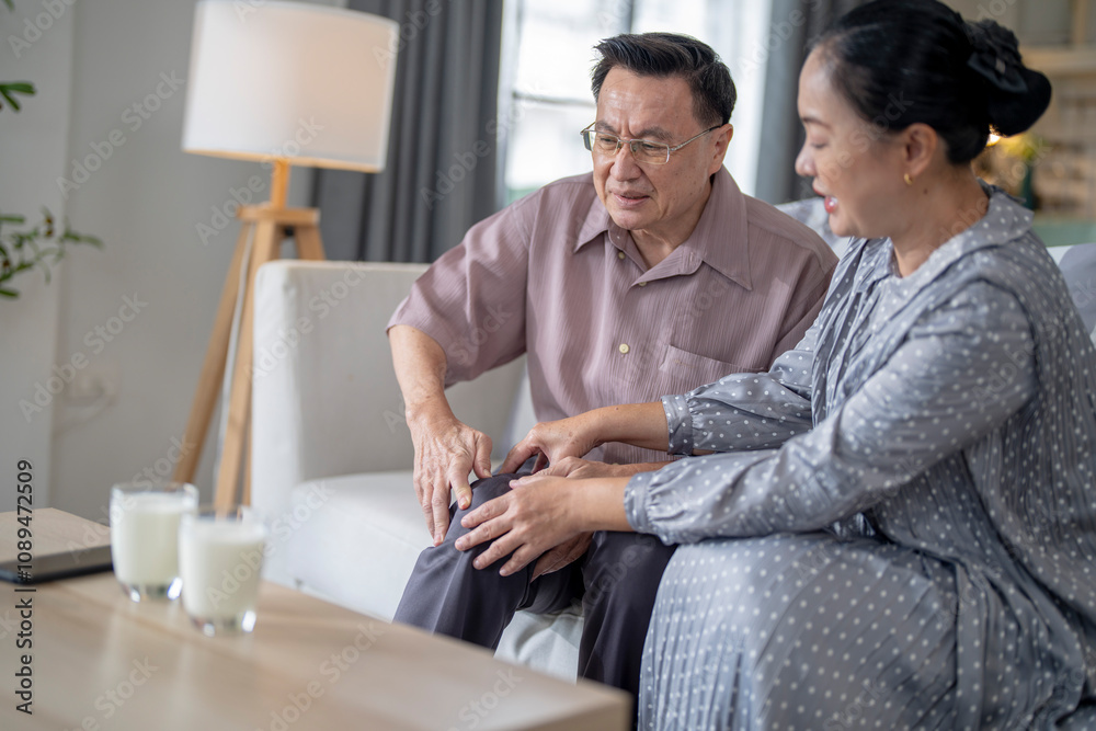 An elderly couple sits in their living room. The man holds his knee, appearing to be in discomfort, while his partner offers support. The image conveys care, concern, and the bond between the couple. An elderly couple sits in their living room. The man holds his knee, appearing to be in discomfort, while his partner offers support. The image conveys care, concern, and the bond between the couple.