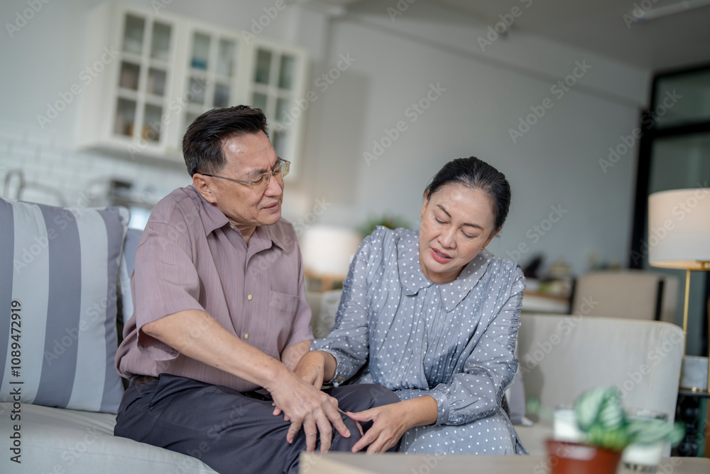 An elderly couple sits in their living room. The man holds his knee, appearing to be in discomfort, while his partner offers support. The image conveys care, concern, and the bond between the couple. An elderly couple sits in their living room. The man holds his knee, appearing to be in discomfort, while his partner offers support. The image conveys care, concern, and the bond between the couple.