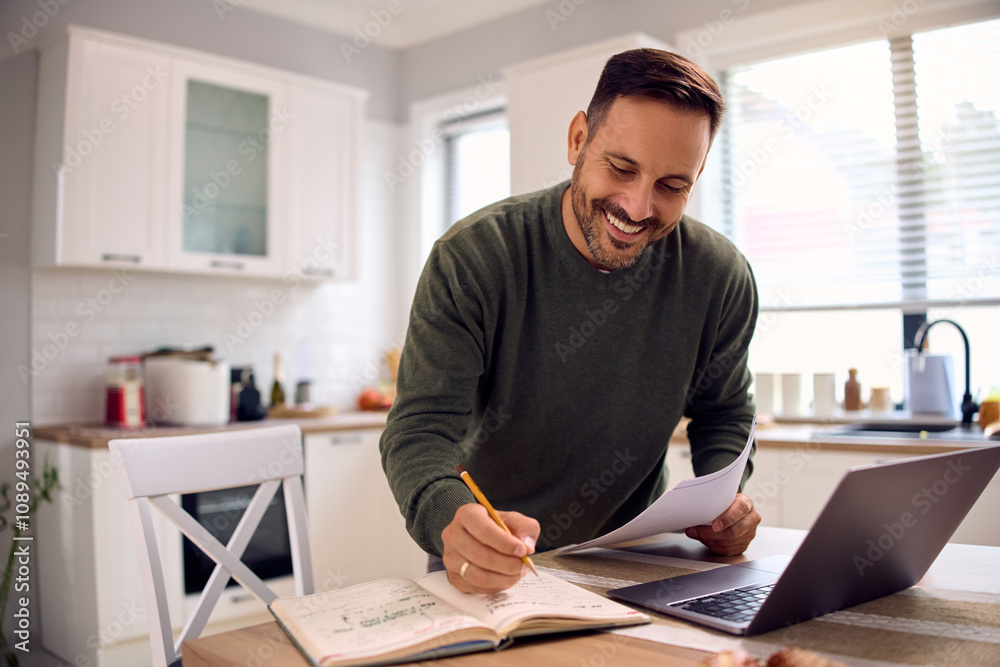 Happy man analysing business reports and taking notes while working on laptop at home Happy man analysing business reports and taking notes while working on laptop at home