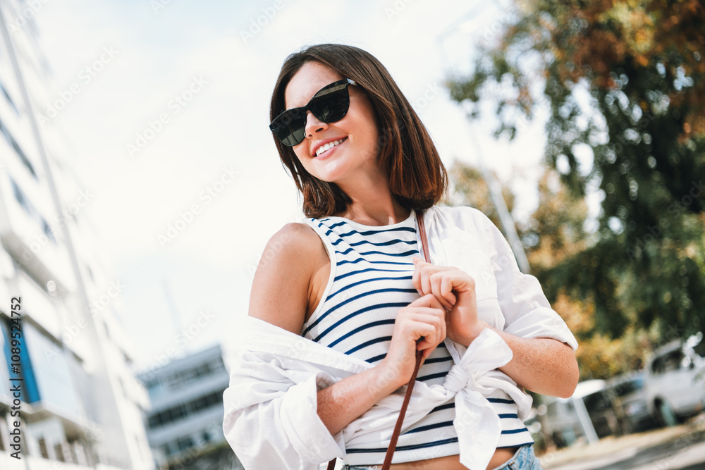 Young woman enjoying a sunny day outdoors, wearing stylish striped shirt and sunglasses in urban setting Young woman enjoying a sunny day outdoors, wearing stylish striped shirt and sunglasses in urban setting