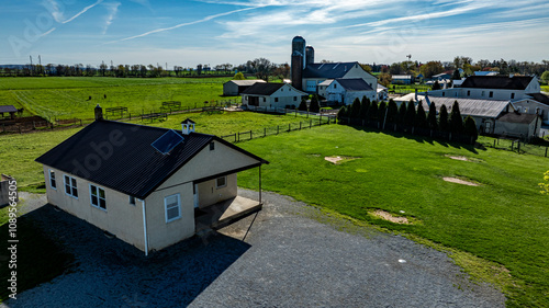 An Amish one room school house sits on a gravel area, surrounded by expansive green fields, barns, and distant structures under a bright blue sky. photo