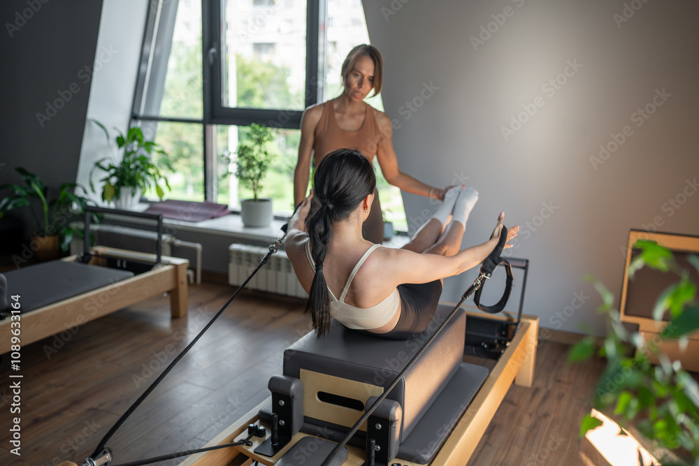 Obraz premium Woman training pilates on the reformer bed with trainer. Reformer pilates studio machine for fitness workouts in gym. Fit, healthy and strong authentical body. Fitness concept