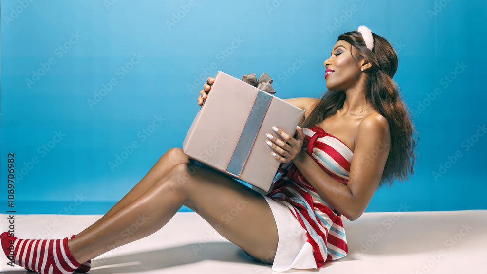 Beautiful seductive black woman sitting and holding Christmas gift, isolated on blue background, festive template, copy space area