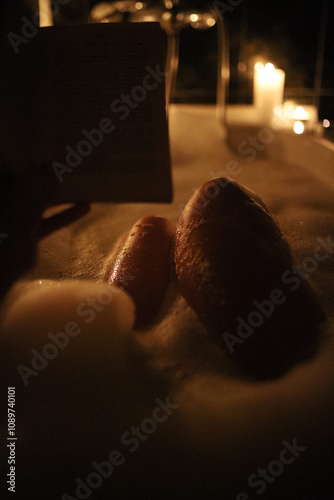Young woman taking bubble bath in a dark bathroom, reading a book and surrounded with candles. Romantic atmosphere