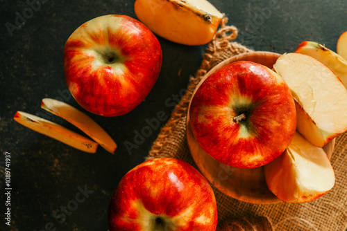 Fresh red apples and apple slices arranged on a rustic burlap cloth with a dark textured background, creating a cozy, autumnal food composition photo