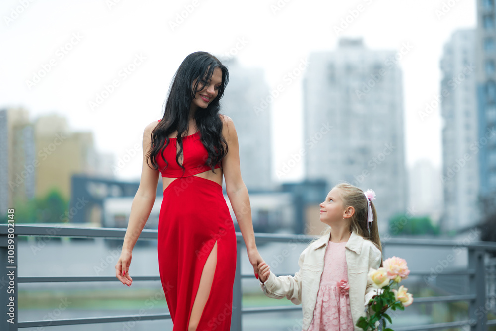Obraz premium Elegant Woman in Red Dress Walking With Young Girl Holding Flowers in City Park