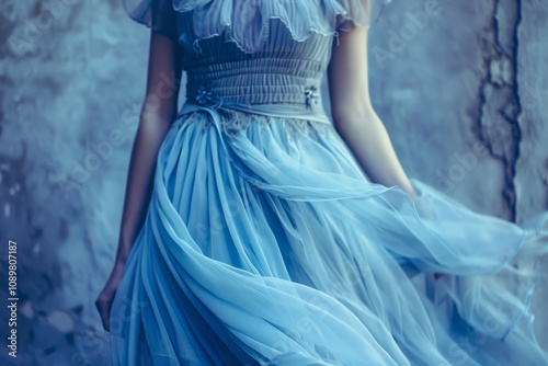 The woman stands elegantly, her delicate blue dress billowing gently around her. The soft fabric contrasts beautifully with the textured gray wall behind her, creating an enchanting mood