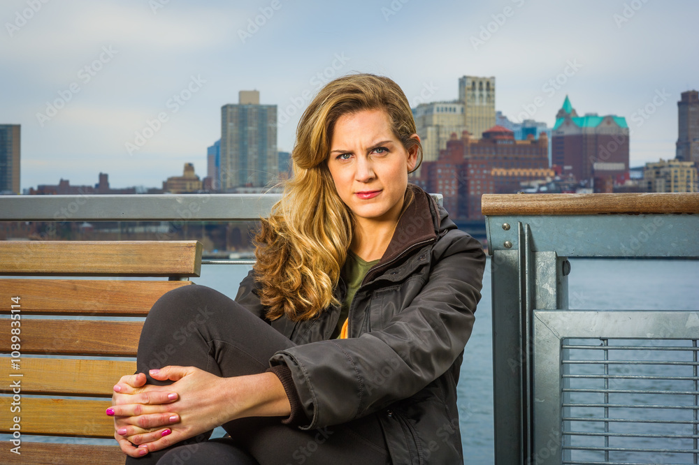 A woman with long blonde hair sits on a wooden bench, wearing a black jacket with the city skyline in the background. The overcast sky creates a muted light over the scene.