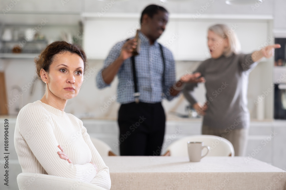 Fototapeta premium Angry middle-aged woman sitting at table while two male and female family members disputing with each other in the back