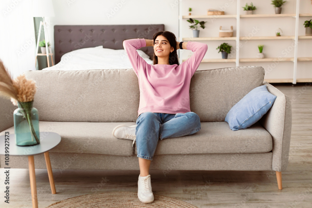 Fototapeta premium Rest Concept. Beautiful lady sitting on comfortable couch at home in living room, looking aside at window. Happy woman relaxing on sofa, leaning back, enjoying weekend free time or break from work